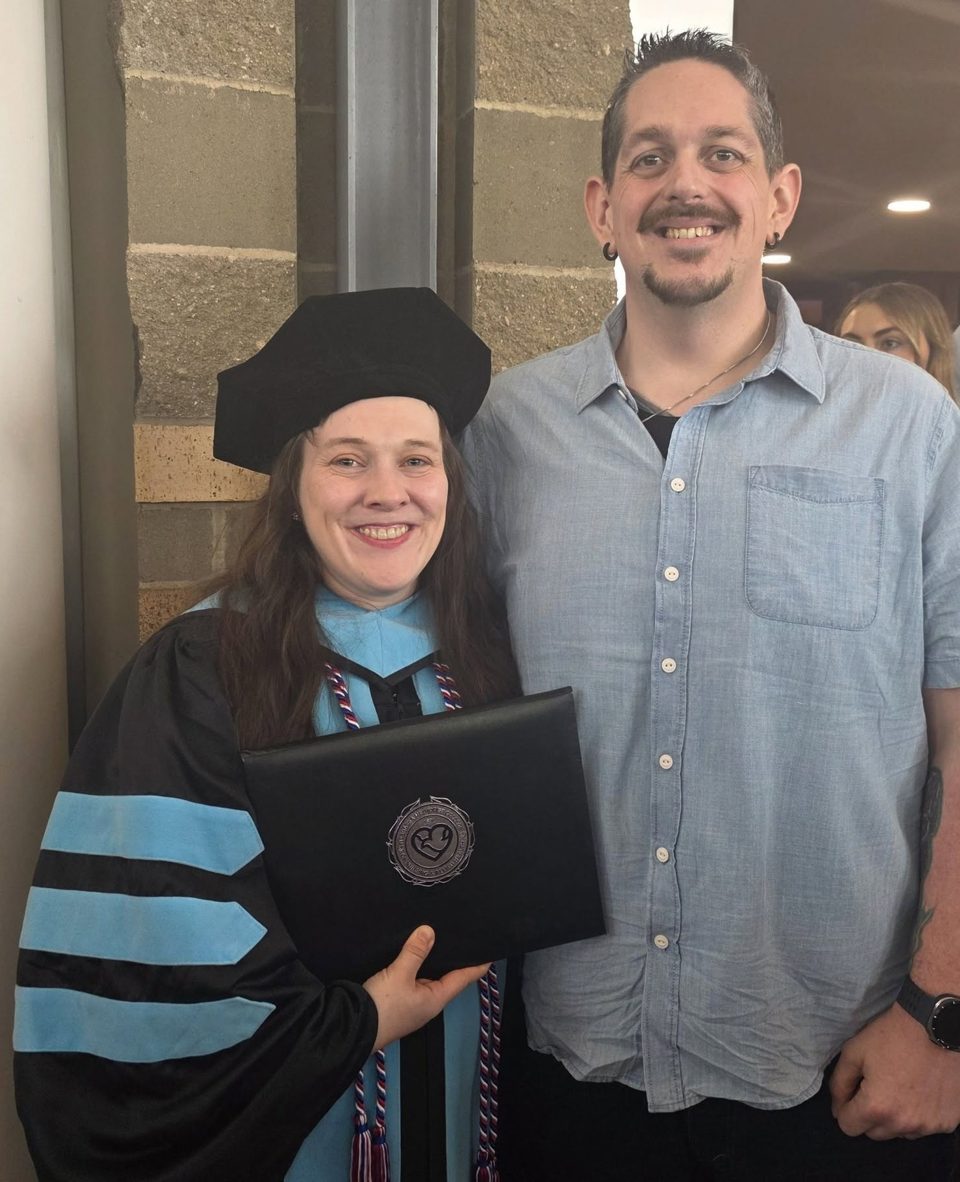 Christina Baynum in doctoral regalia holding her diploma, standing with Thomas Baynum